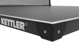 Close-up of a Kettler table tennis table showing the black side panel with the white KETTLER logo, the dark playing surface, and part of the net against a white background.