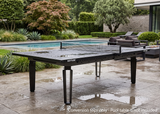 A black Kettler outdoor pool table with a net, positioned on a wet stone patio next to a swimming pool in a landscaped garden.